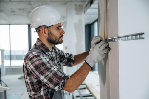 Ein Bauarbeiter mit Schutzhelm und Handschuhen misst eine Wand in einem renovierten Innenraum.